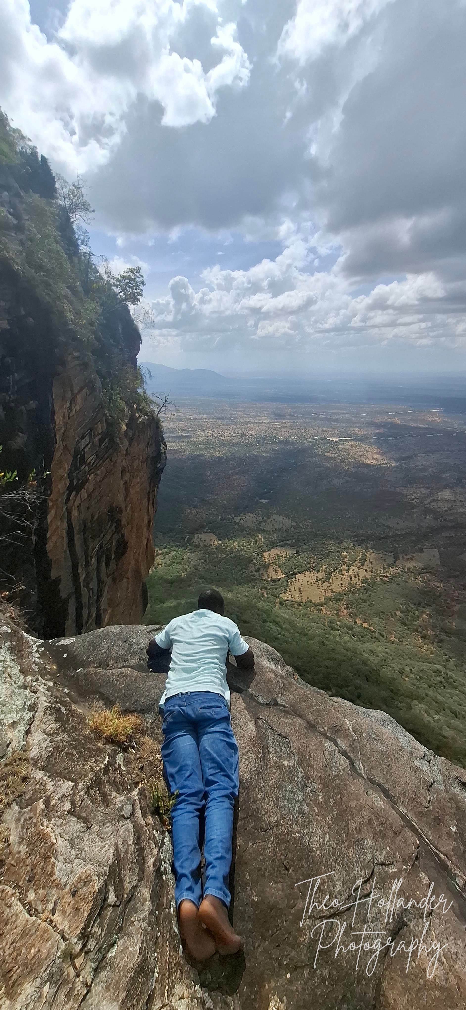 A traveller lying at the edge of a sheer cliff, looking out over the valley below